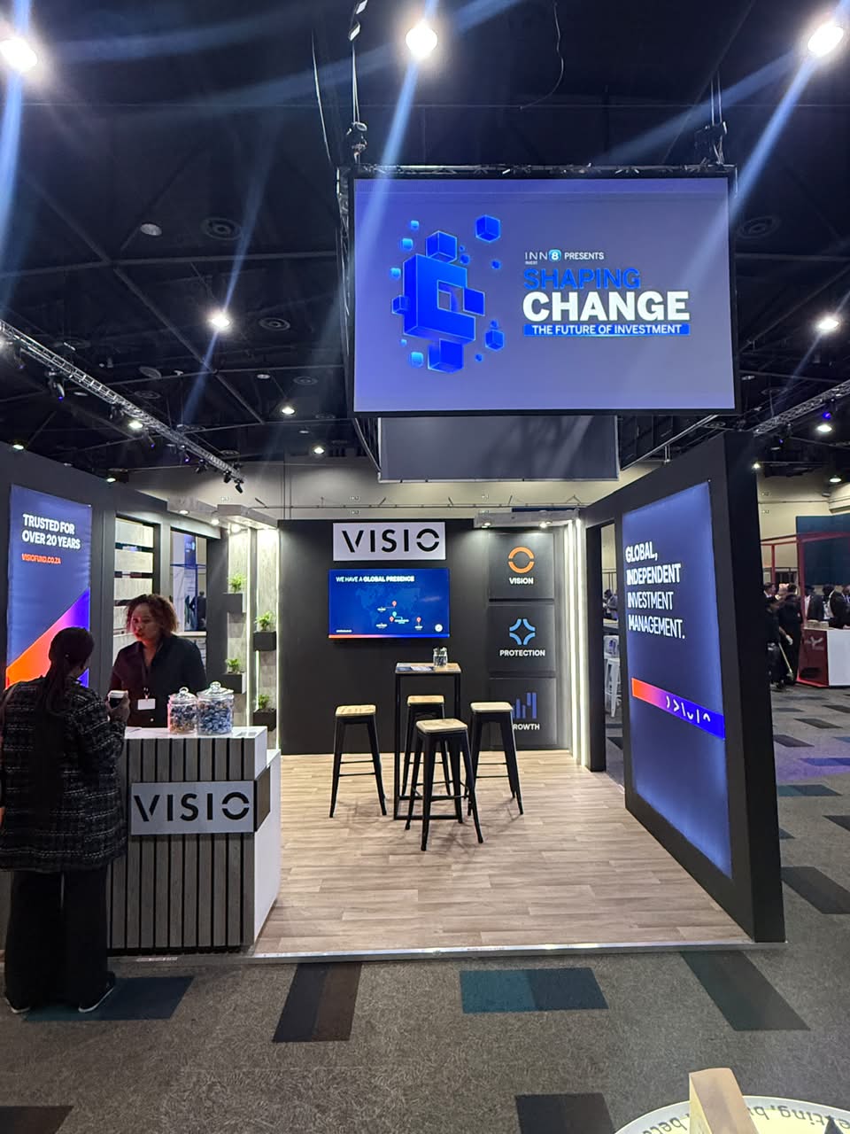 Custom exhibition stand built for Visio, with backlit wood-slat panelling, lounge seating and 'Global, Independent Investment Management' messaging
