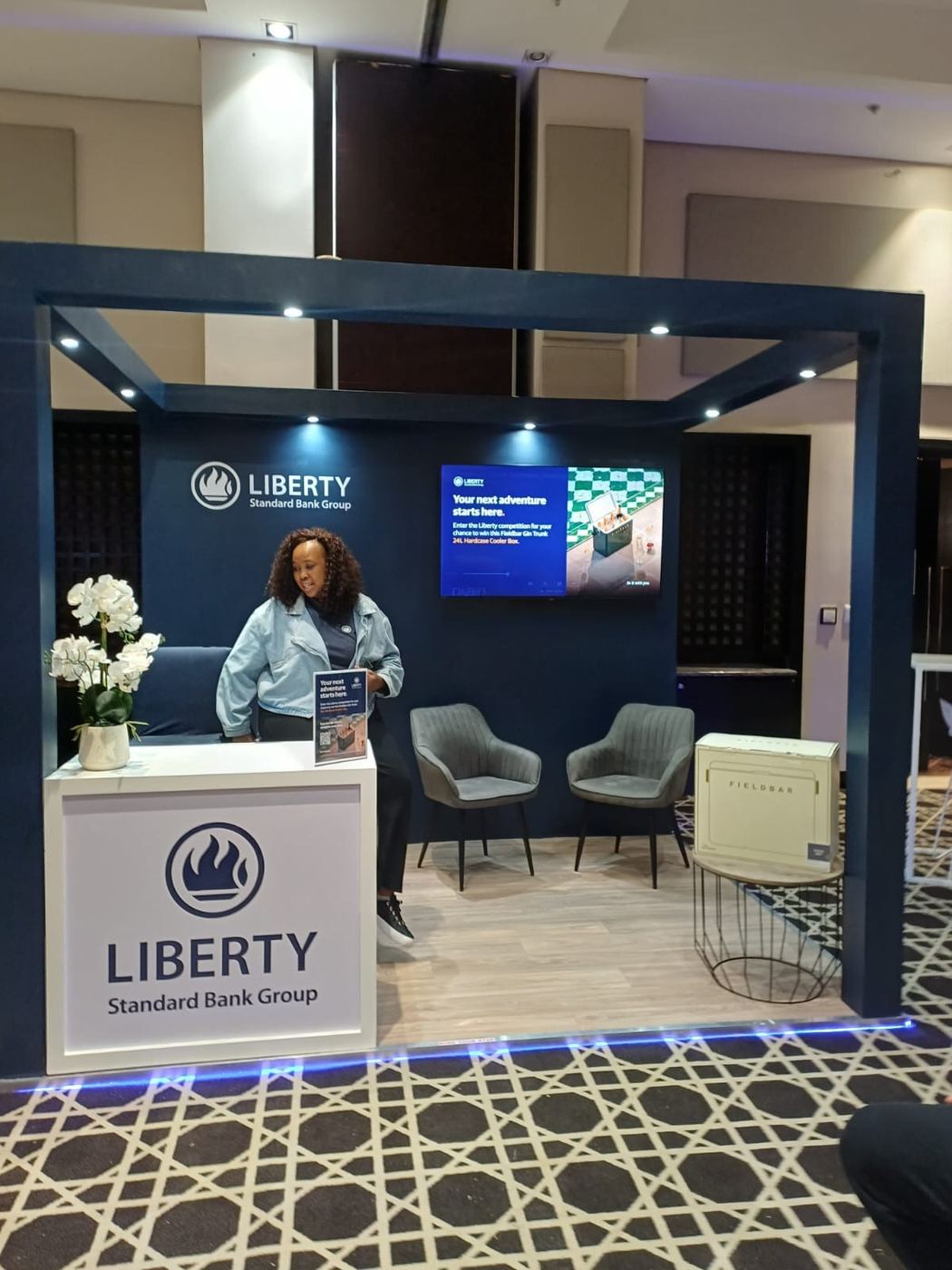Custom exhibition stand built for Liberty, Standard Bank Group, with a navy reception desk, lounge seating and a digital promotional screen