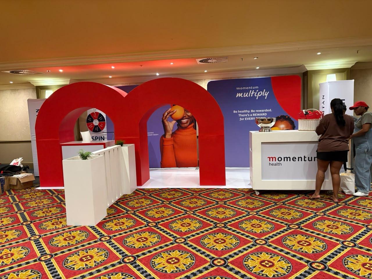 Custom exhibition stand built for Momentum Health, with a bold red archway entrance, a spin-wheel game and 'Momentum Multiply' branding