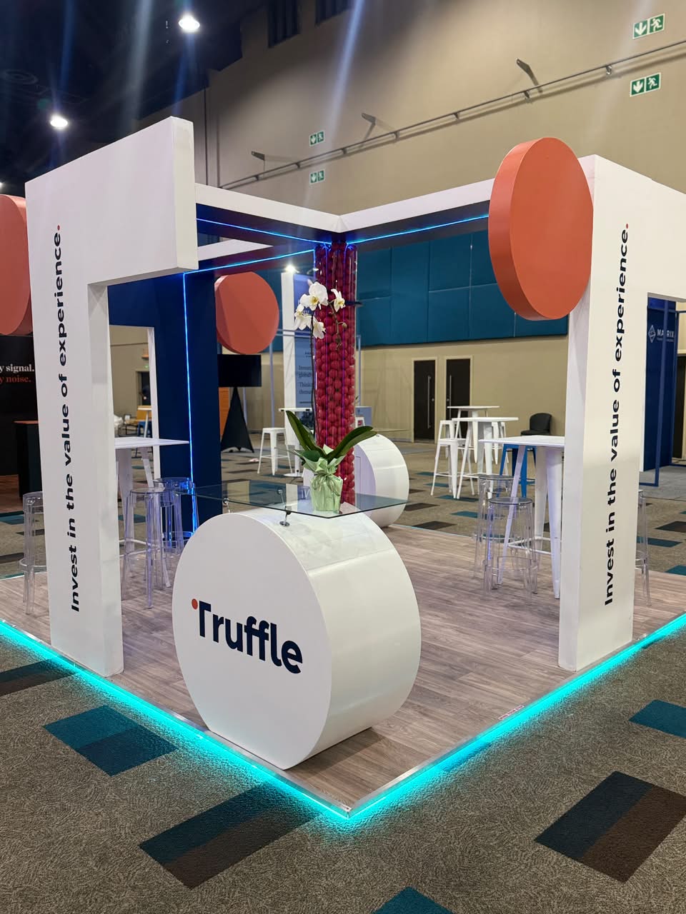 Custom exhibition stand built for Truffle, with a glass-top reception table, orange circular accents and 'Invest in the value of experience' branded messaging