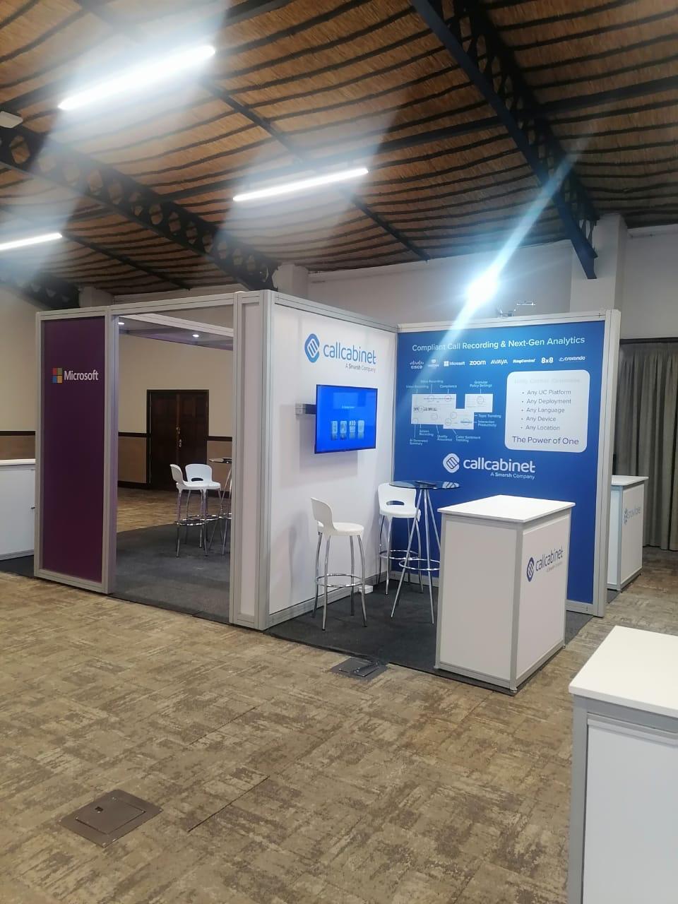Shell scheme exhibition stand built for CallCabinet, with a wall-mounted screen, branded backdrop and an adjoining Microsoft co-branded meeting nook